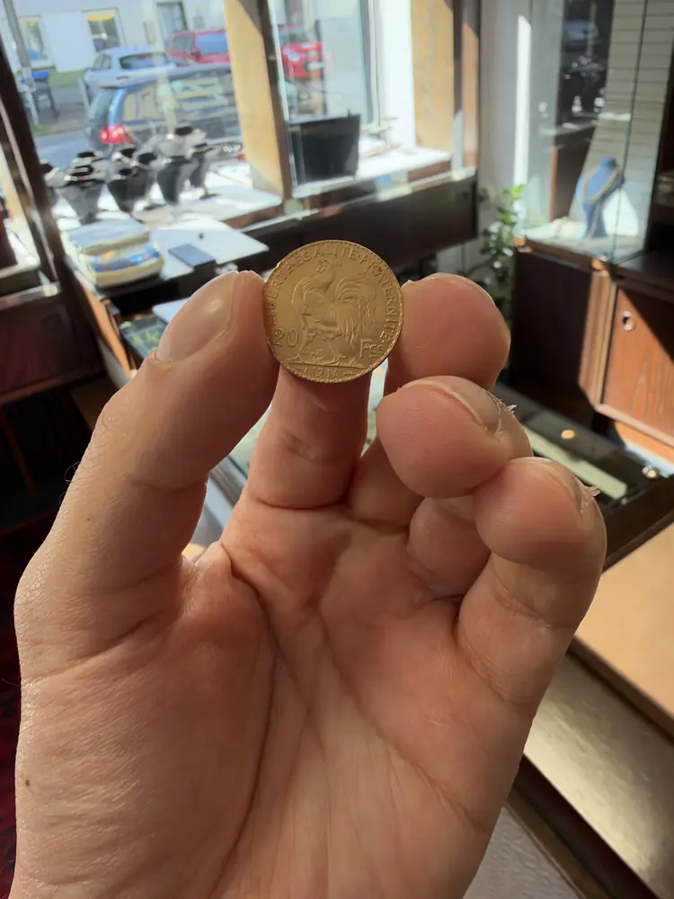Hand hält eine französische Goldmünze mit Hahnmotiv vor einer Ladentheke