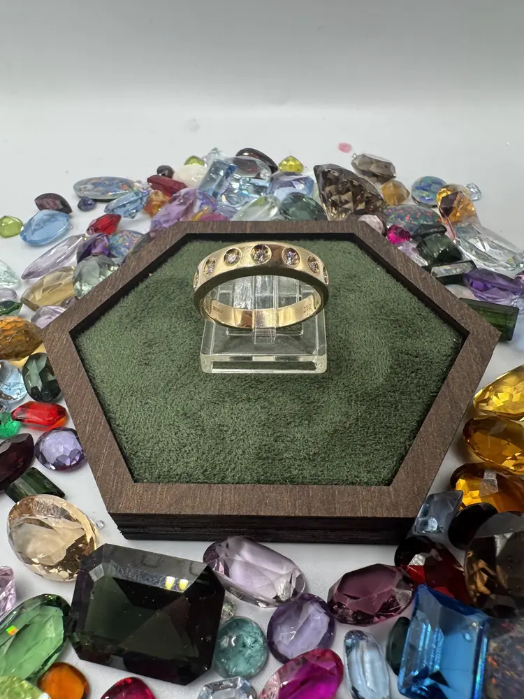 Goldener Bandring mit eingelassenen Steinen auf einem Ringhalter, umgeben von bunten Edelsteinen